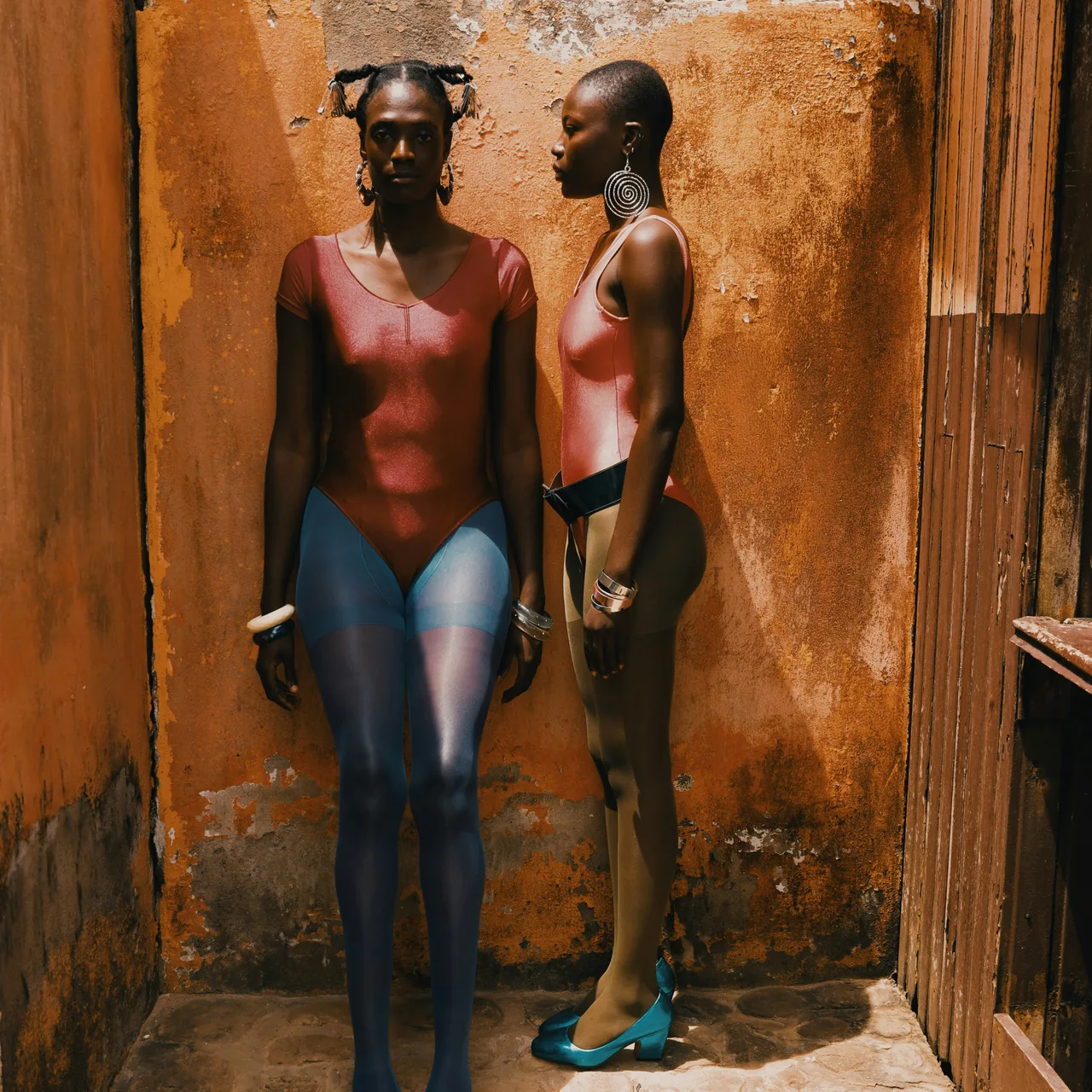 Two women wearing red tops standing against a sunlit orange wall