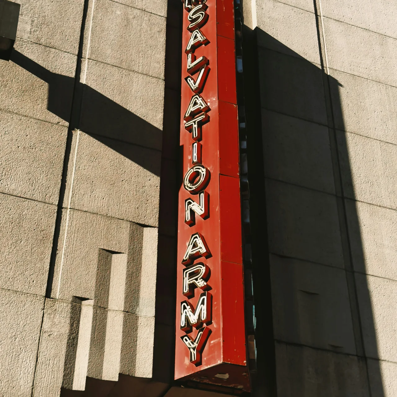 A vertical red Salvation Army sign casting a long shadow across a stone facade