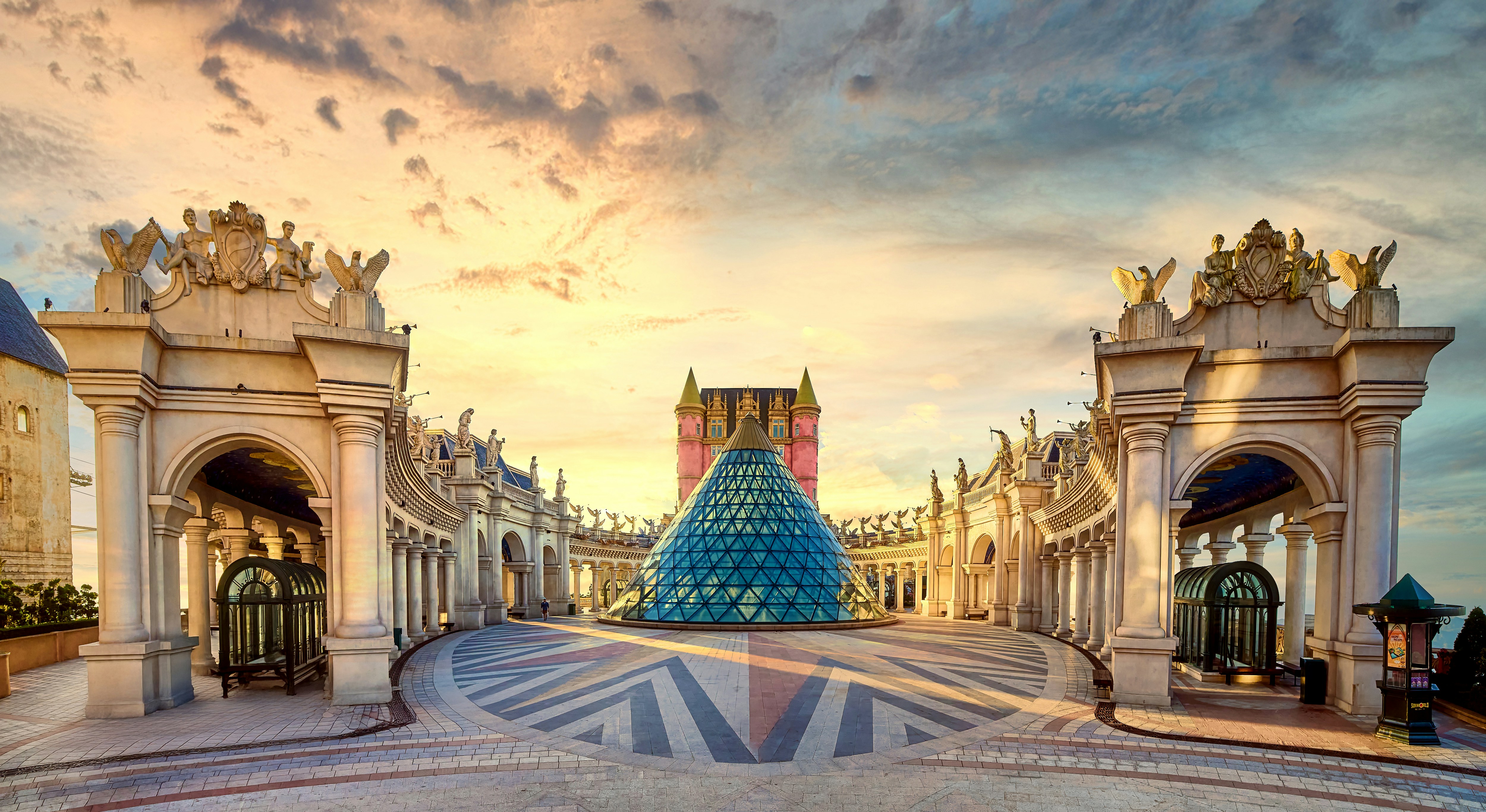 European-style architectural plaza with a glass pyramid and grand colonnades under a golden sunset sky.