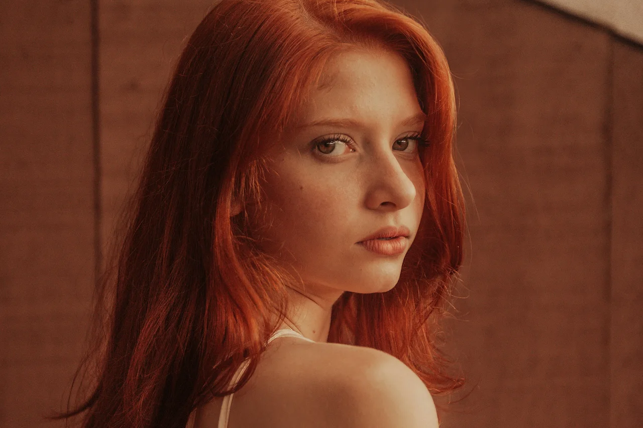 A red-haired woman looking over her shoulder beside a warm brick wall