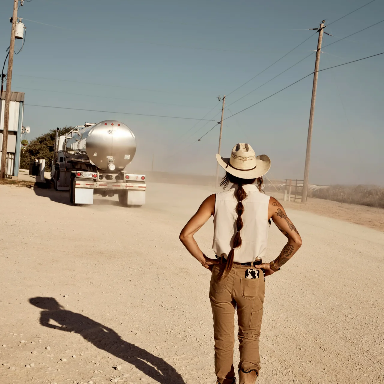 A person in a cowboy hat watching a tanker truck on a dusty sunlit road