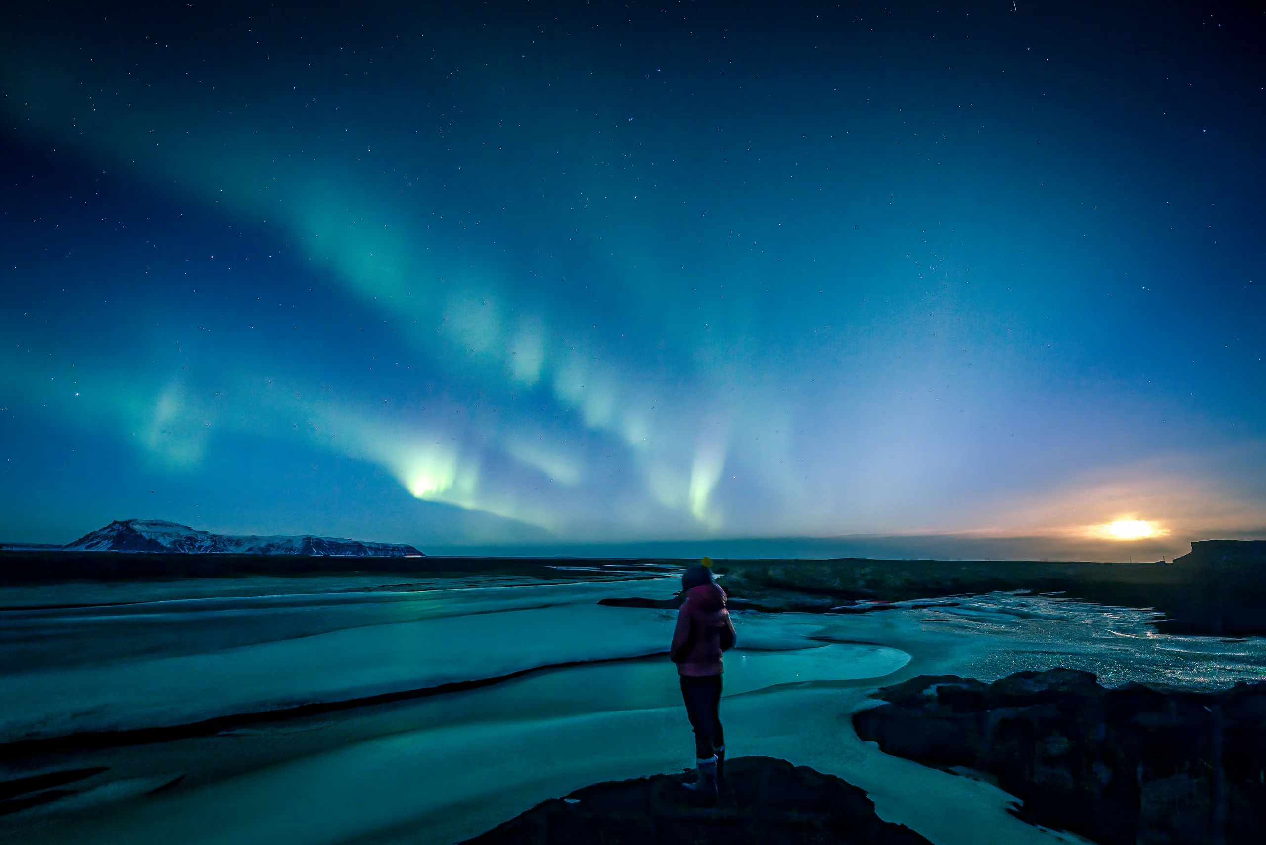 Lone figure under a green aurora over still water
