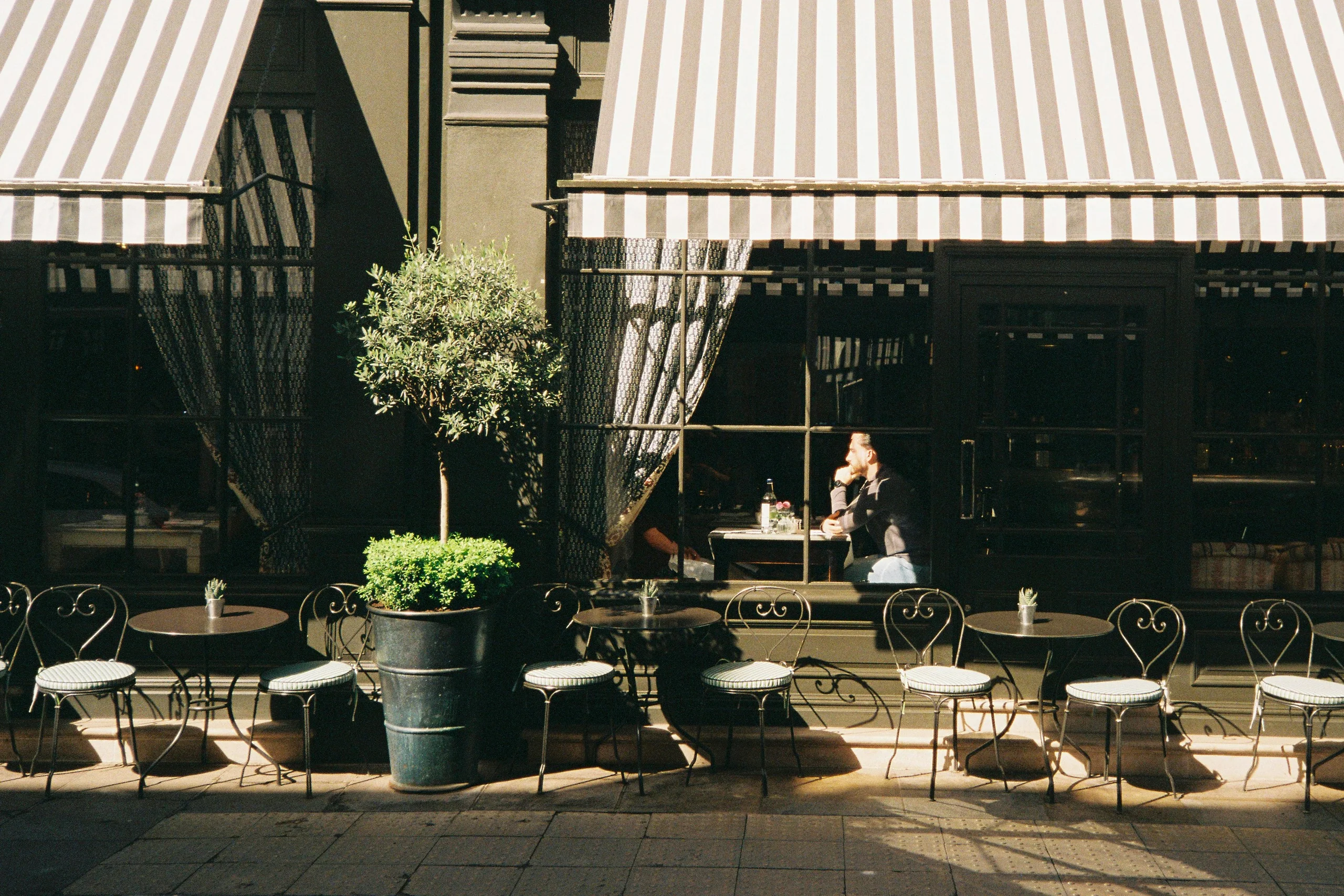 A sunlit café terrace with striped awnings and one guest inside