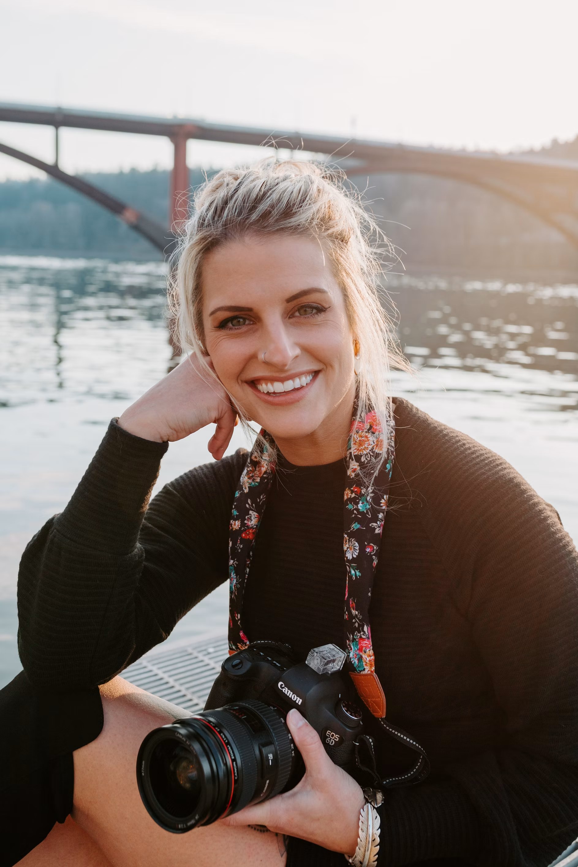 Isabelle Fontaine smiling with a camera in her hands on a riverbank in late light, a bridge behind her