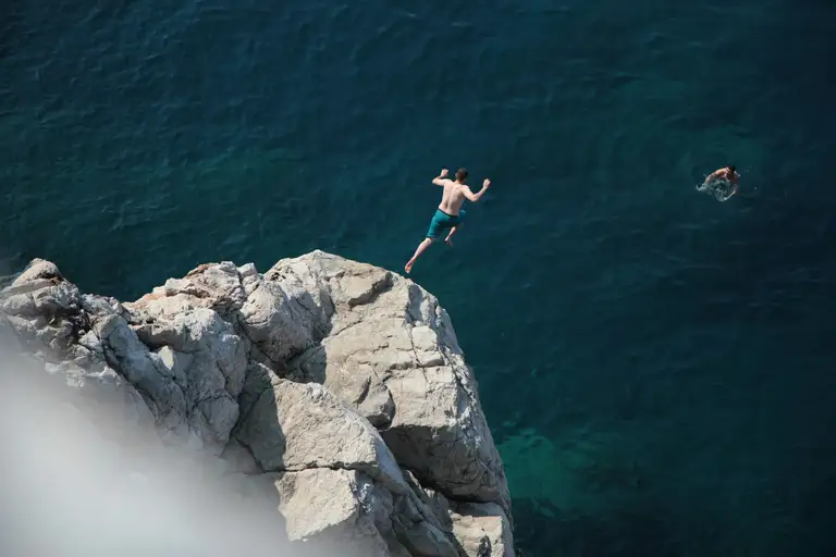 Small figure leaping from a rocky cliff into turquoise sea water